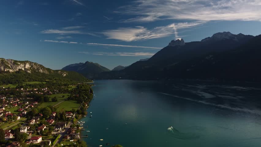High aerial footage of a shot looking up Lake Annecy in early morning sunshine. Drone moves sideways to the right starting at the shore line over houses and fields then moving out over the water.