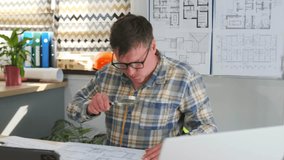 caucasian architect studying blueprints at desk, laptop open, tablet nearby, measuring tape and helmet on table, potted plant, pinned floorplans, focused expression, adjusting layout details - Powered by Shutterstock - Get 15% off with code: PIKWIZARD15