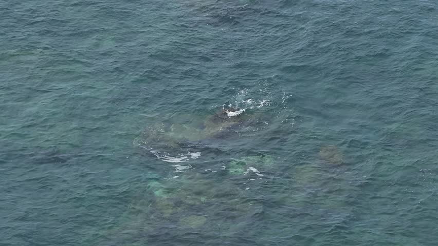 Aerial view over small rock submerged reef with ocean waves washing over making white foam