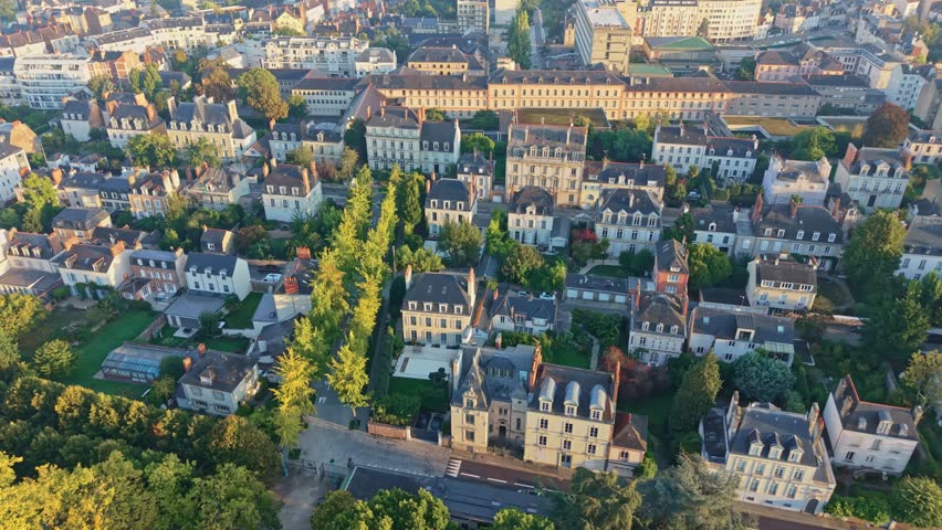 Drone shot moving backward above Thabor Park in Rennes, showing houses, trees, and the city under warm sunrise light.