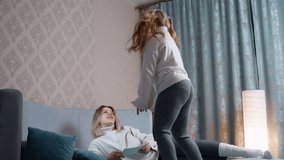 Cheerful mother relaxes on bed holding notebook while playful daughter jumps energetically nearby, creating dynamic scene of joy, fun, and family bonding in cozy bright bedroom with curtains - Powered by Shutterstock - Get 15% off with code: PIKWIZARD15