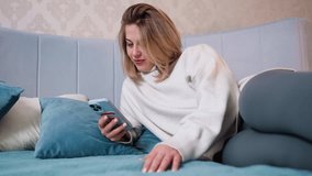 Woman relaxing on bed using smartphone, dressed in cozy white sweater and gray leggings, leaning on soft cushions, comfortable home environment with pastel tones, showing leisure, modern technology - Powered by Shutterstock - Get 15% off with code: PIKWIZARD15