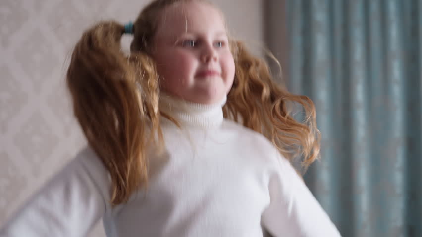 Joyful young girl with long blonde hair tied in pigtails wearing white turtleneck sweater smiles brightly while jumping playfully indoors, expressing carefree childhood happiness,fun in cozy home
