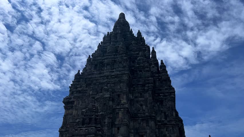 View of the ancient Prambanan Temple, a Hindu temple complex in Yogyakarta, Indonesia. The temple rises above the green foliage under a blue sky. The towers cast shadows on a sunny day. 4К