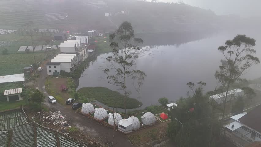 Aerial view of a foggy highland lake near Sikunir, Dieng Plateau, showing lakeside houses, terraced farmland, and misty mountain surroundings in Central Java, Indonesia