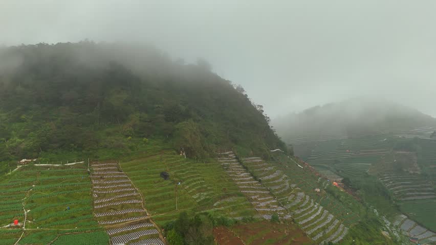Aerial rotate view of a calm highland lake near Sikunir, Dieng Plateau, showing lakeside houses, terraced farmland, and misty mountain surroundings in Central Java, Indonesia