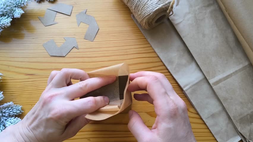 Gift wrapping made from eco-friendly materials against a background of a recycling symbol and Christmas tree branches. Fold the edges of the packaging