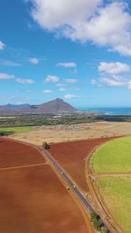 Vertical aerial shot of a road on Mauritius island landscape with coastal lagoons, towns, and distant mountains under dramatic tropical clouds and sunlight.