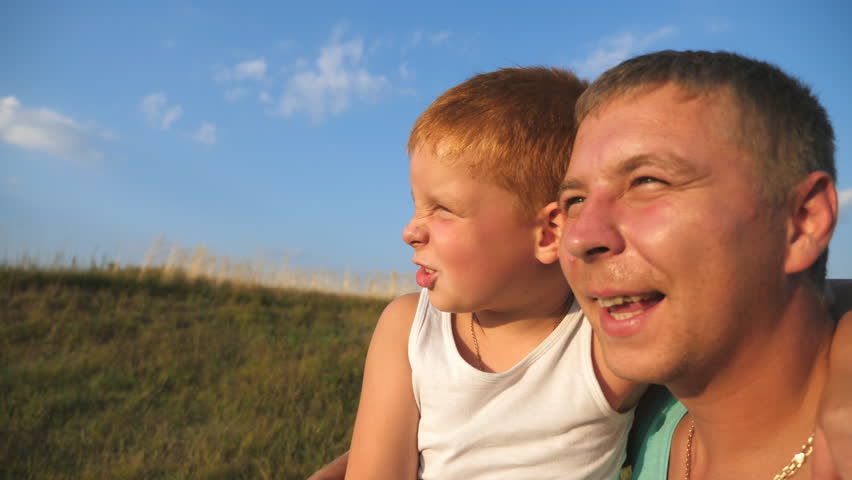 Close up of red-haired boy sitting at green grass on the lawn with his father and pointing at something to him. Dad and his little son relax at meadow. Happy family spending time together at nature