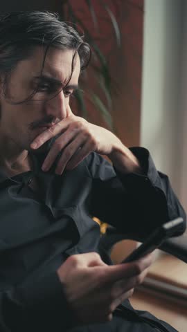 In contemplation, businessman close-up of a handsome young white European man with Slavic features, long hair, mustache, and expressive eyes, deeply engaged with his smartphone in warm lighting.