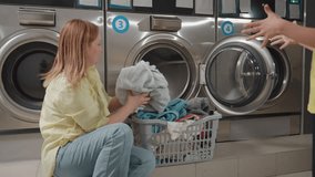 Mother with toddler packing clothes into washer at laundromat, woman kneels by open stainless machine, child gestures while basket of towels waits, sorting textiles, clean service routine inside - Powered by Shutterstock - Get 15% off with code: PIKWIZARD15