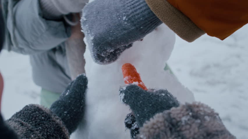 Closeup of parents and child using gloved hands to place carrot nose on snowman in snowy forest