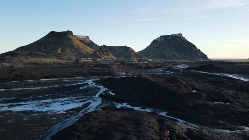 Aerial view of stark black sand contrasting with winding rivers and snow-capped mountains under a pale sky, creating a dramatic landscape, Vik, Myrdalshreppur, Iceland.