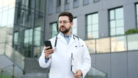 Confident doctor in white coat using mobile phone while standing outside near hospital building. Handsome male medical worker physician checks messages, browses web or chats online with patient - Powered by Shutterstock - Get 15% off with code: PIKWIZARD15