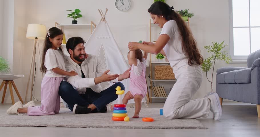 Happy parents guide toddler steps at home. In living room, caring parents and child girl support a smiling baby learning to stand near toys and a teepee. Family bonding, first stepping and leisure.