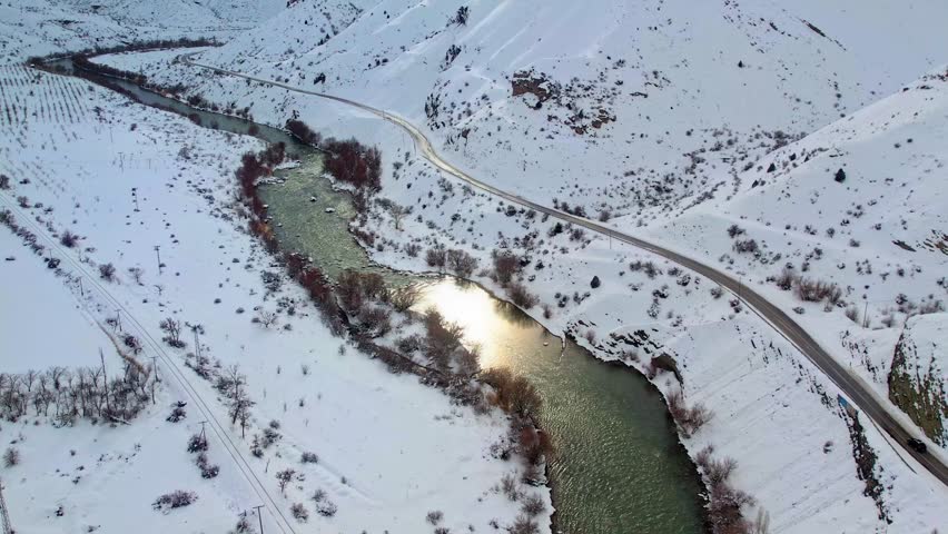 A winding river flows through a snowy valley as a mountain road follows its edge. Leafless winter trees and white hills create a calm, natural winter scene.