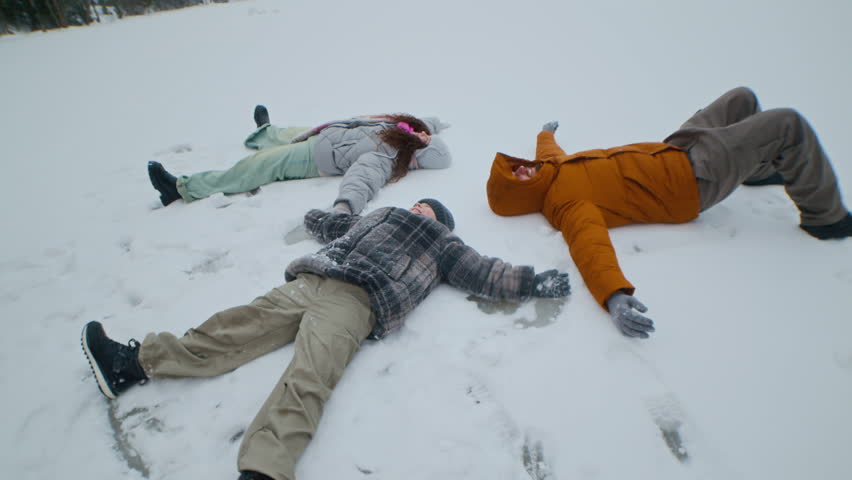 Family of three lying on snowy ground with arms and legs outstretched making snow angels together in open winter field