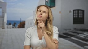 Blonde woman thinking in a sunny city street setting with mediterranean architecture and ocean view in the background, expressing deep thought and contemplation outdoors. - Powered by Shutterstock - Get 15% off with code: PIKWIZARD15