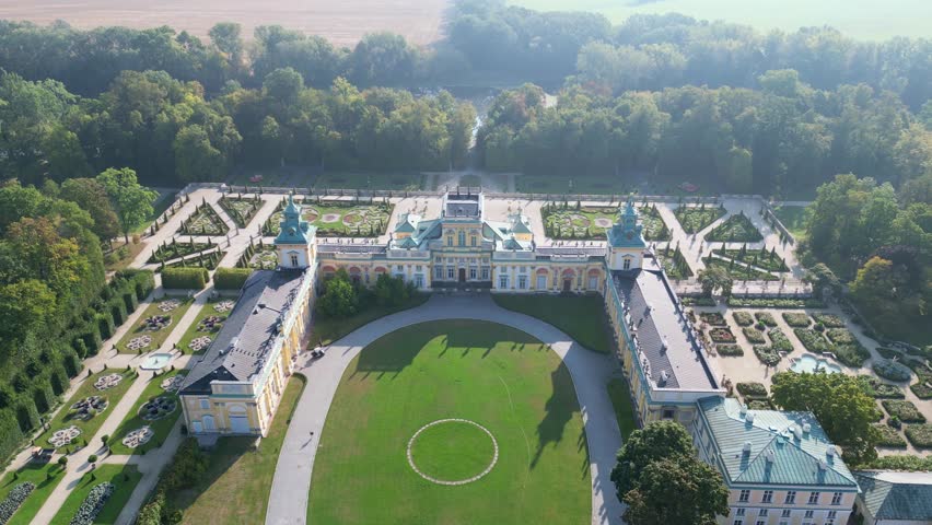Stunning drone view of Wilanów Palace in Warsaw with ornate baroque facade, colorful roof, fountains and vast landscaped gardens bathed in morning light.