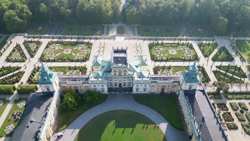 Stunning drone view of Wilanów Palace in Warsaw with ornate baroque facade, blue colorful roof, and vast landscaped gardens bathed in morning light.
