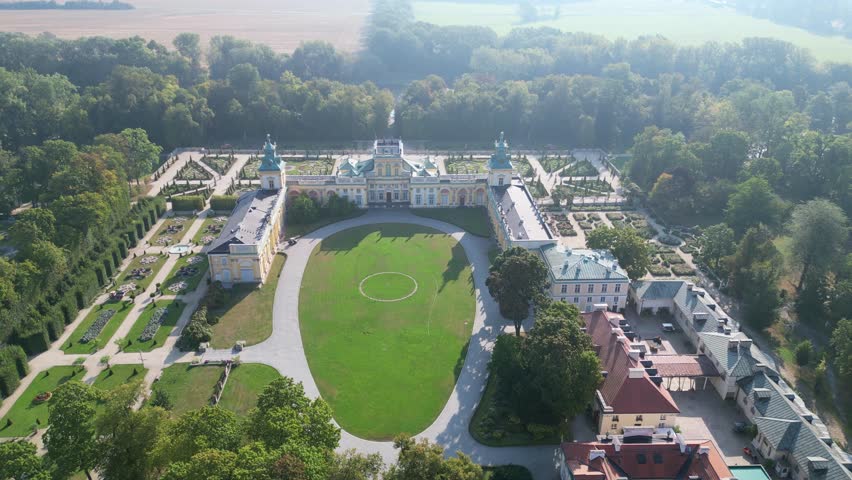 Stunning drone aerial view of Wilanów Palace in Warsaw with ornate baroque facade, colorful roof, and vast landscaped gardens bathed in morning light.