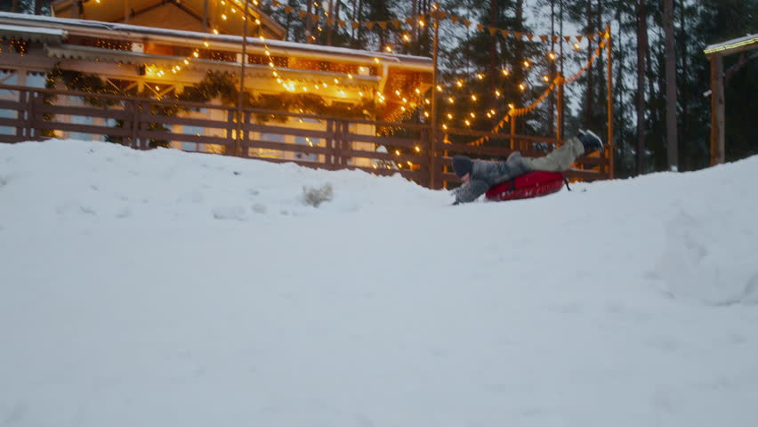 Child lying on inflatable sled sliding down snowy slope in winter park with warm glowing lights in background