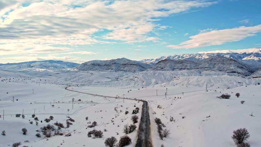 Aerial view of a winding mountain road cutting through a vast snow-covered landscape. Winter hills and sunlit mountains create a dramatic and peaceful scene ideal for travel, nature, and cinematic pro