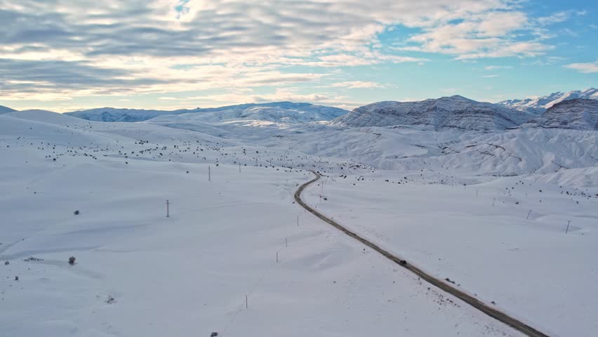 Aerial view of a winding mountain road cutting through a vast snow-covered landscape. Winter hills and sunlit mountains create a dramatic and peaceful scene ideal for travel, nature, and cinematic pro