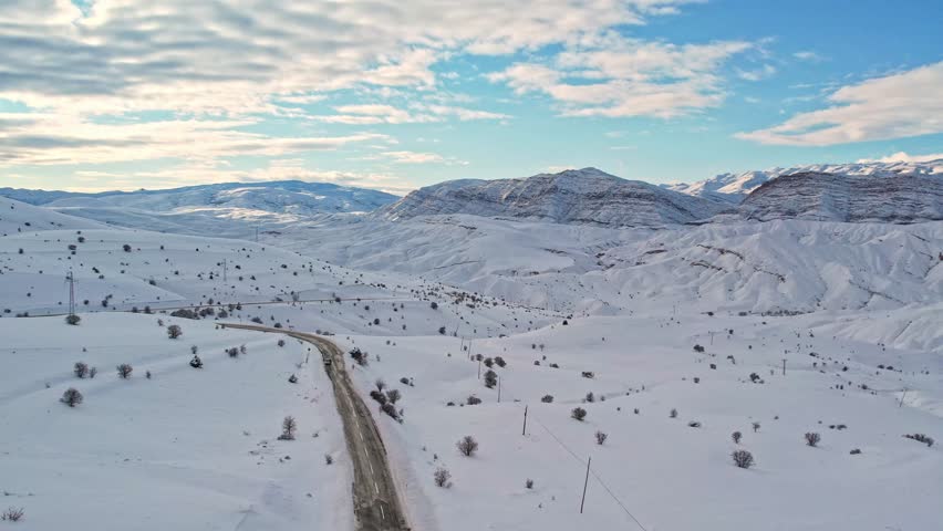 Aerial view of a winding mountain road cutting through a vast snow-covered landscape. Winter hills and sunlit mountains create a dramatic and peaceful scene ideal for travel, nature, and cinematic pro