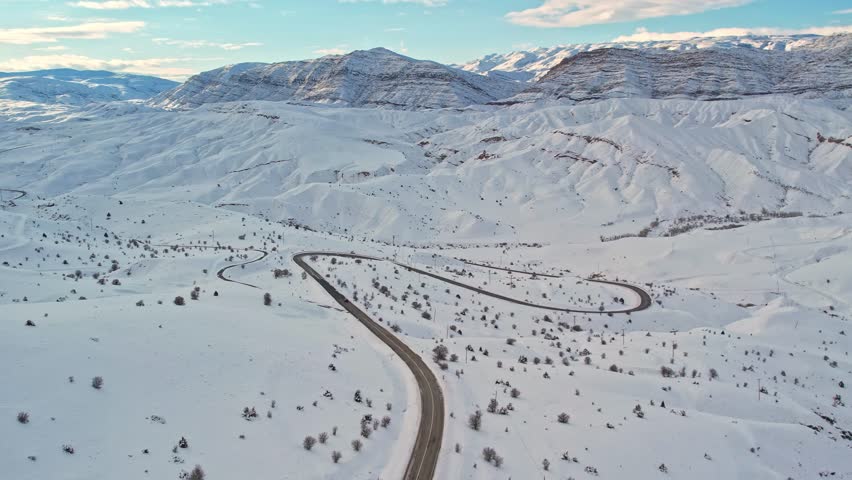 Aerial view of a winding mountain road cutting through a vast snow-covered landscape. Winter hills and sunlit mountains create a dramatic and peaceful scene ideal for travel, nature, and cinematic pro