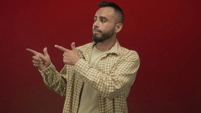 Hispanic young man pointing fingers with both hands in red studio wearing checked shirt and beige t shirt, slight shrug and neutral expression; casual skepticism. - Powered by Shutterstock - Get 15% off with code: PIKWIZARD15