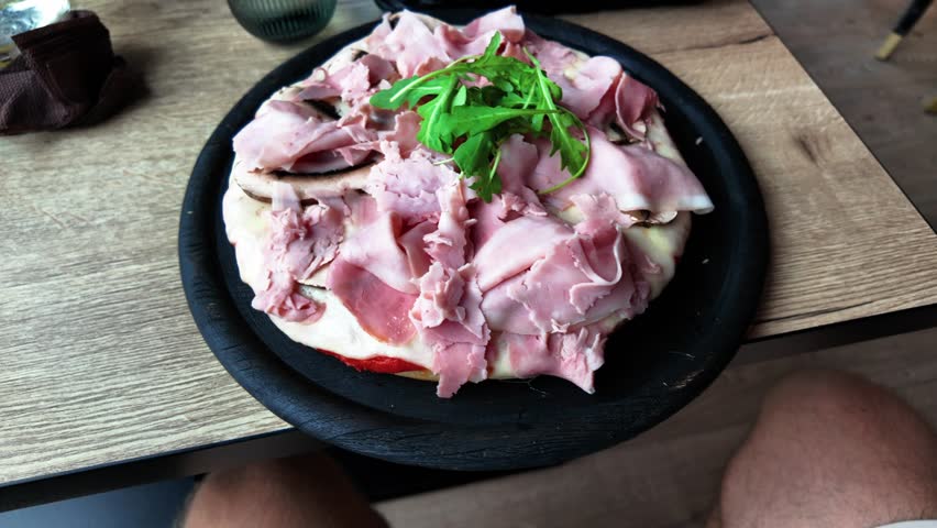 Freshly prepared pizza topped with slices of ham and arugula, served on a black plate over a wooden table in a cozy dining setting. Ideal for food and culinary themes.