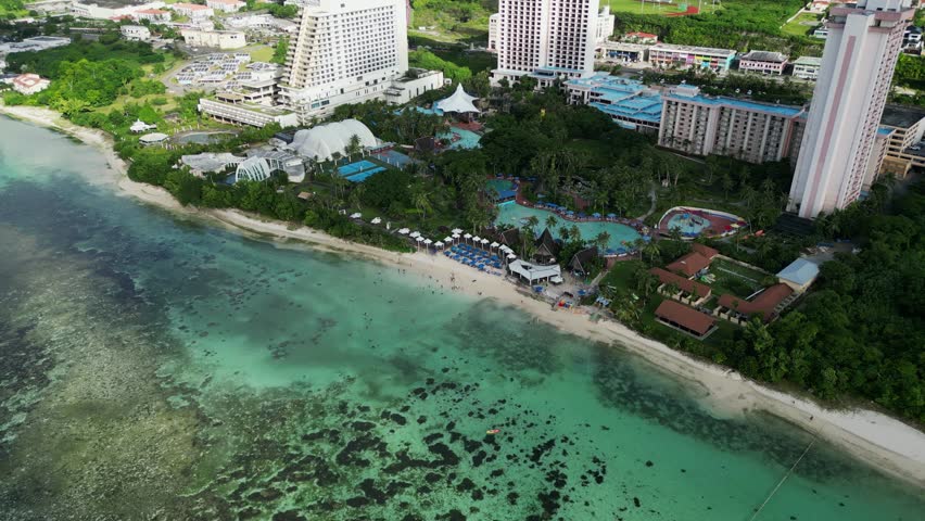 Picturesque aerial flyover shot of turquoise ocean coastline and hotel resort amid lush greenery at Guam, USA during daytime