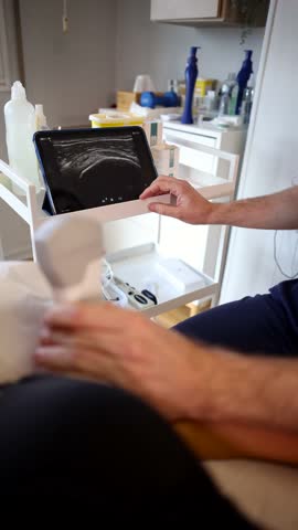 Experienced medical professional conducting a musculoskeletal ultrasound examination on a patient's knee joint, using a portable probe and analyzing the real time image on a digital tablet screen