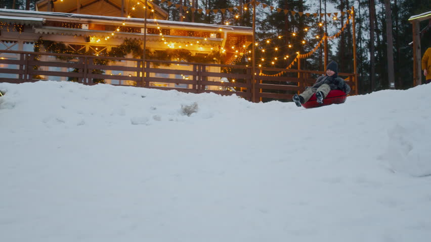 Boy sliding downhill on inflatable sled across snowy terrain with decorative winter lights in background