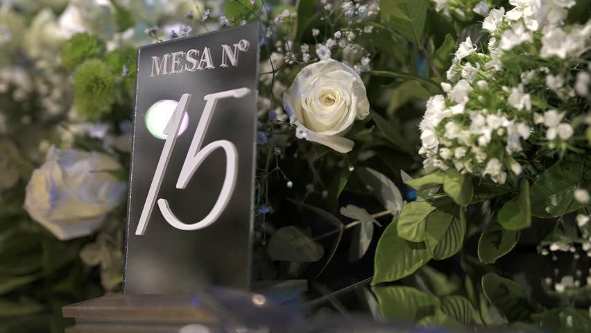 A close-up of an elegant wedding table centerpiece, featuring a modern black and white "MESA Nº 15" sign surrounded by lush white roses and greenery.