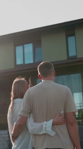 Young couple embracing in their backyard, gazing at their modern suburban home. The new homeowners are seen from behind, looking ahead to their shared future with excitement and love