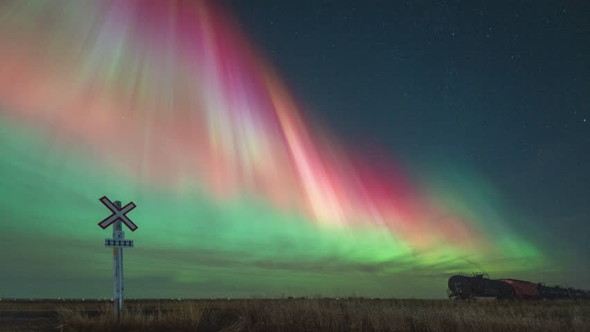 Colourful northern lights (Aurora Borealis) dance over parked train cars at a crossing in the prairie fields near Tuxford, Saskatchewan, Canada.