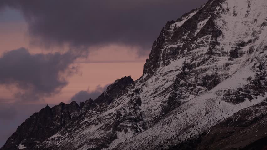A dramatic close-up of the rugged, snow-covered mountains of Torres del Paine, Chile. Features a striking contrast against a purple-pink sky at dawn or dusk, emphasizing the cold and majestic scale.