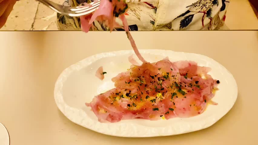 plate containing thinly sliced cured meat, possibly high-quality ham, prosciutto, or carpaccio. The slices are lightly dressed with olive oil and sprinkled with chopped herbs (like parsley).