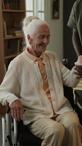 Vertical shot of white haired elderly woman moving from wheelchair to sofa at home with unrecognizable African American man as social worker assisting carefully
