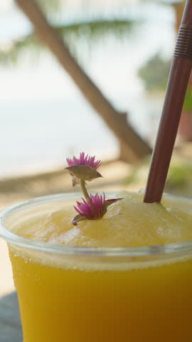 Frozen Mango Smoothie in Plastic Cup with Straw and Flowers on Beachfront Table Tropical Drink Refreshment.