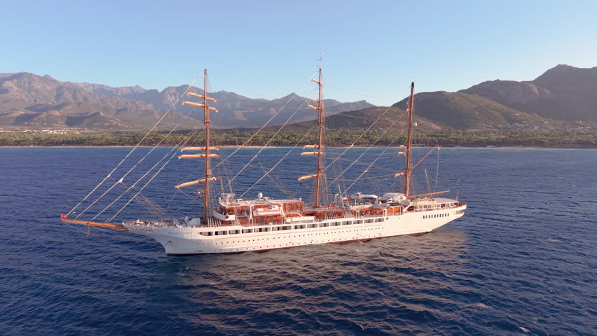 Aerial elegant threemasted sailing ship cruising at dawn by rocky island shores amid tranquil blue seas. Calvi. Corsica. France