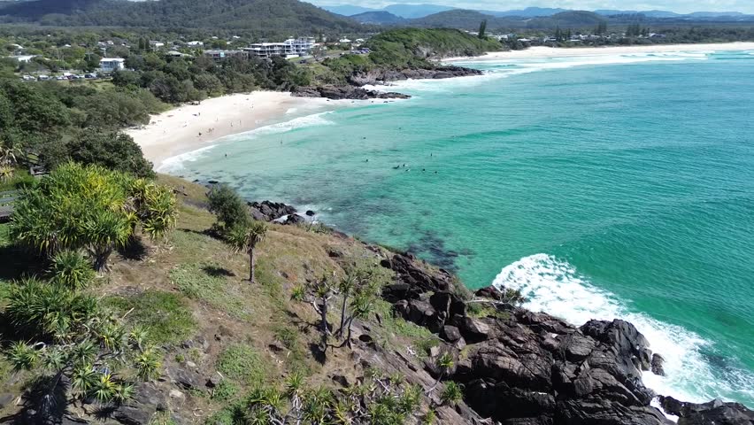 A drone flying over a cliff and group of people surfing on the sea behind it near the beach