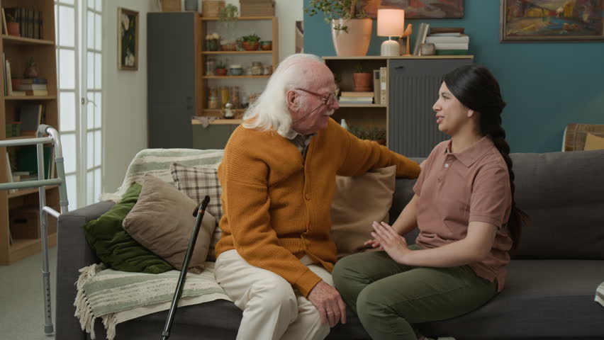 Front view shot of young woman embracing white haired senior man while sitting on couch at home and enjoying conversation