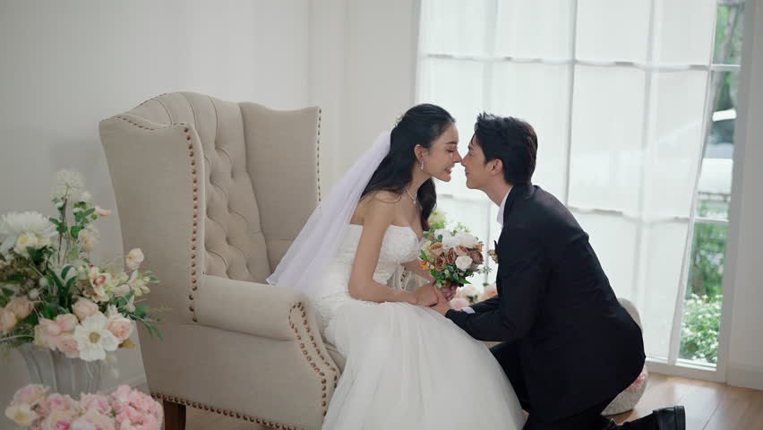 Asian Groom Kneeling and Kissing Brides on wedding day, Happy Asian couple in wedding attire, Bride wearing a beautiful white wedding dress and groom in a classic suit.