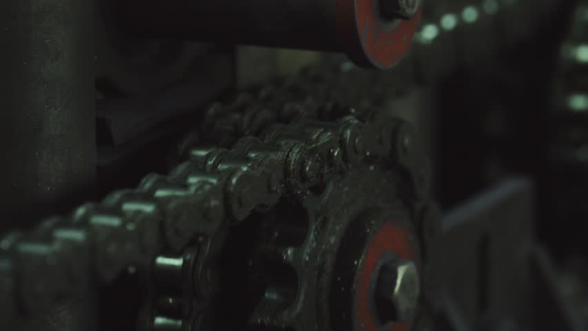 Close up of a dirty, oily roller chain turning on a sprocket gear as part of a large industrial machine, illustrating mechanical power transmission and industrial manufacturing concepts