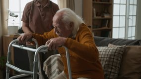 Side view shot of white haired senior man getting up from couch using walker with shaking hands, young woman as female social worker assisting - Powered by Shutterstock - Get 15% off with code: PIKWIZARD15