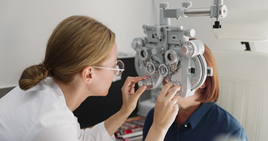 Young optometrist woman conducting vision examination for elderly female patient using professional phoropter in modern ophthalmology clinic. Medical eye diagnostics and eye care services, close up