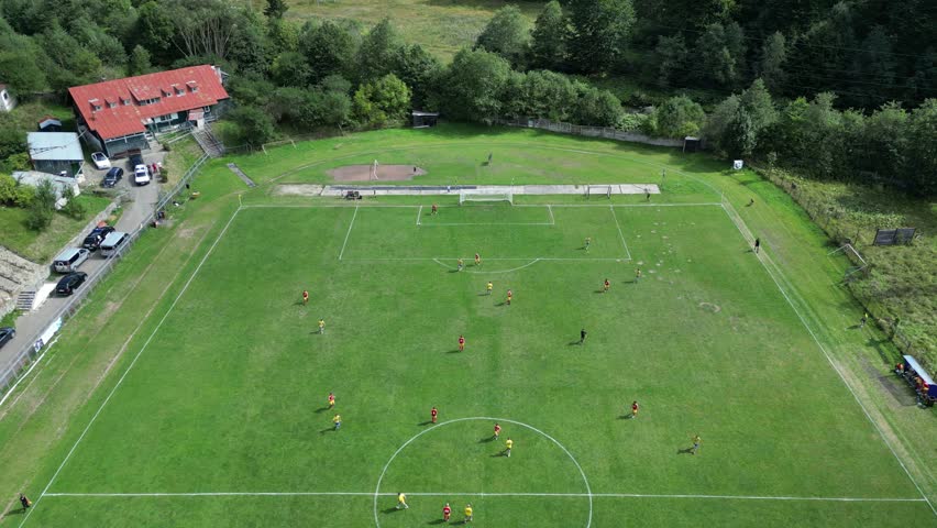 An aerial drone shot of a football pitch during the match between two teams. A small group of fans can be noticed on the tribune.